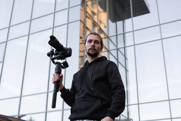 istock Young Professional videographer holding professional camera on 3-axis gimbal stabilizer. Pro equipment helps to make high quality video without shaking. Cameraman wearing white hoodie making a videos. 1216134088