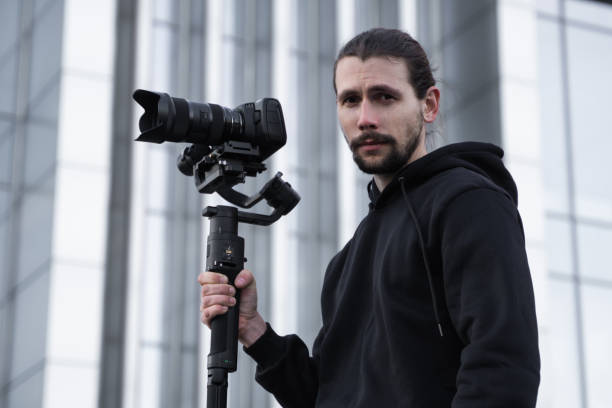 istock Young Professional videographer holding professional camera on 3-axis gimbal stabilizer. Pro equipment helps to make high quality video without shaking. Cameraman wearing white hoodie making a videos. 1216134780