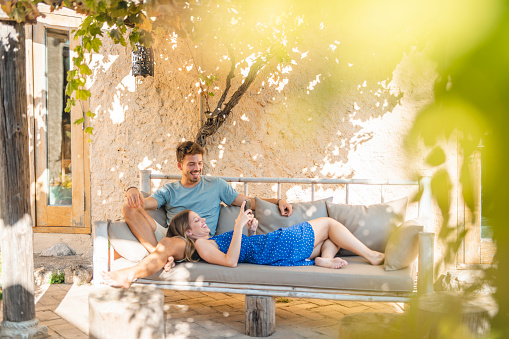 Close-up of Spanish male and female couple in mid 20s relaxing outdoors on sofa under shady arbor and checking smart phone.
