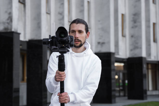 istock Young Professional videographer holding professional camera on 3-axis gimbal stabilizer. Pro equipment helps to make high quality video without shaking. Cameraman wearing white hoodie making a videos. 1216135262