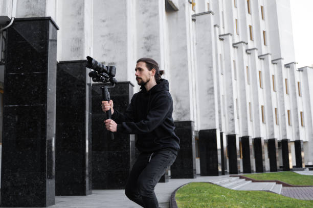 istock Young Professional videographer holding professional camera on 3-axis gimbal stabilizer. Pro equipment helps to make high quality video without shaking. Cameraman wearing black hoodie making a videos. 1216135468