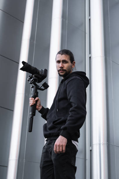 istock Young Professional videographer holding professional camera on 3-axis gimbal stabilizer. Pro equipment helps to make high quality video without shaking. Cameraman wearing white hoodie making a videos. 1216137477