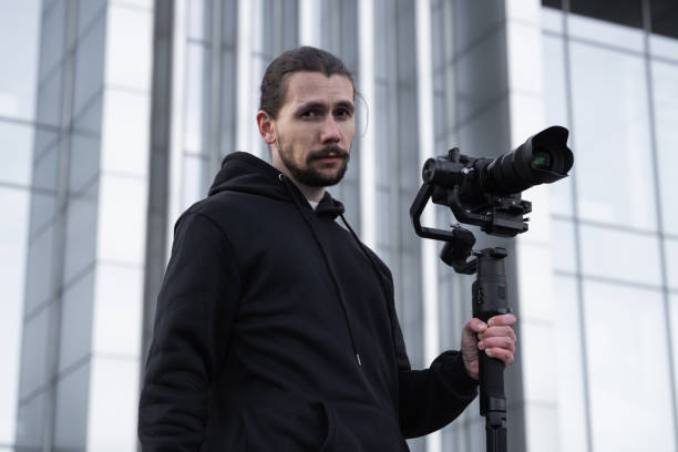 istock Young Professional videographer holding professional camera on 3-axis gimbal stabilizer. Pro equipment helps to make high quality video without shaking. Cameraman wearing white hoodie making a videos. 1216137658