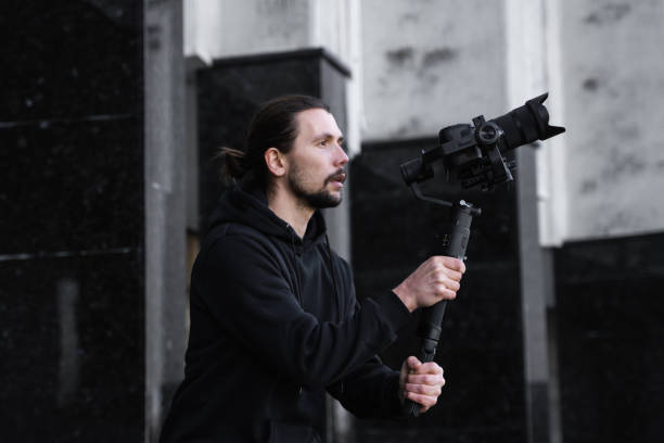 istock Young Professional videographer holding professional camera on 3-axis gimbal stabilizer. Pro equipment helps to make high quality video without shaking. Cameraman wearing black hoodie making a videos. 1216137718