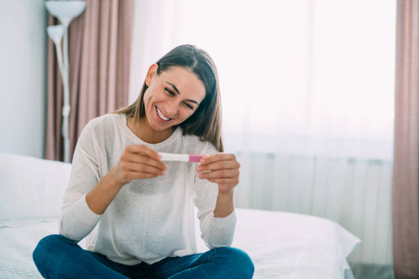 Happy pregnant woman. Cheerful young woman sitting on the bed and looking at pregnancy test. Happy young woman holding in hands positive pregnancy test in bedroom. in vitro fertilization photos stock pictures, royalty-free photos & images