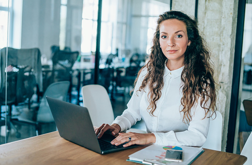 Portrait of beautiful businesswoman sitting at her desk in office. Confident woman working on laptop and looking at camera.