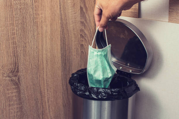 Disposable medical masks in a hand let down to infectious waste trash bin.