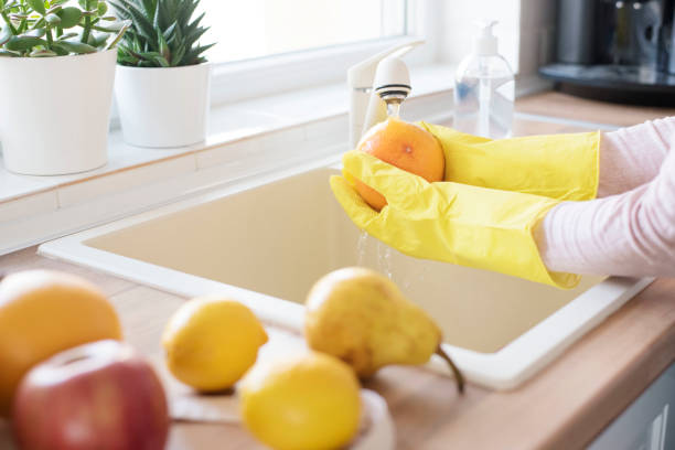 donna con guanti protettivi che lava frutta fresca a casa in cucina, detersivo in primo piano. concetto di igiene durante l'epidemia di coronavirus - contemporary domestic room sparse indoors foto e immagini stock