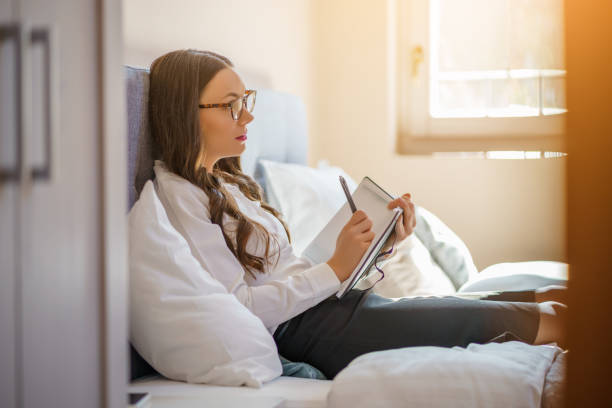 Woman is at home by herself in her apartment in the city. She is writing in her journal diary, a great way to contemplate life and mental wellbeing.