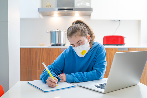 Blond teen girl doing homework at home in Coronavirus quarantine with mask notebook and computer at kitchen