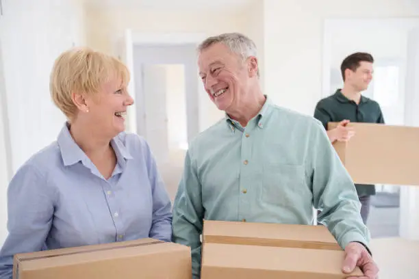 Photo of Senior Couple Downsizing In Retirement Carrying Boxes Into New Home On Moving Day With Removal Man Helping