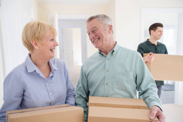 senior couple downsizing in retirement carrying boxes into new home on moving day with removal man helping - downsizing imagens e fotografias de stock