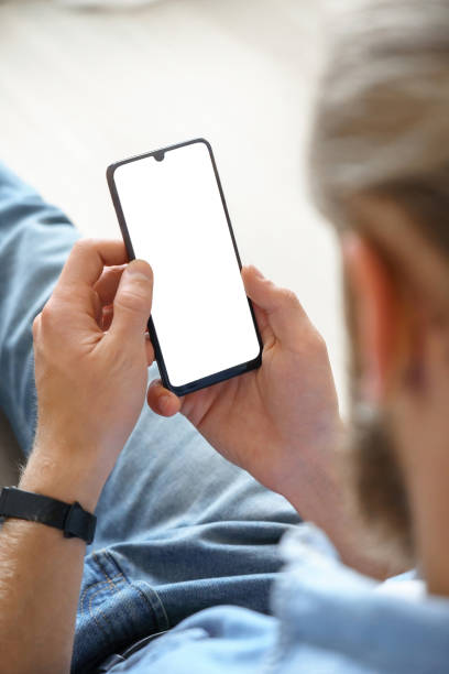 istock Millennial man user holding smart phone mock up white screen in hand using mobile dating app, check social media, order food delivery, reading news sit on couch at home. Over shoulder vertical view. 1216158101