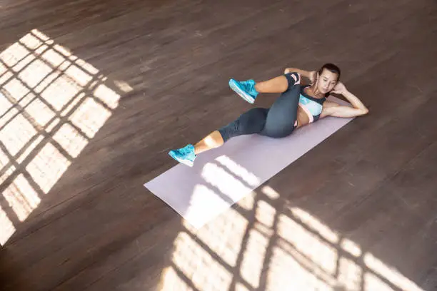 Photo of Sporty fit active young woman doing bicycle crunch situp exercise alone lying on mat wooden floor, strong sportswoman wear activewear training abs core muscle workout in sport gym studio, top view