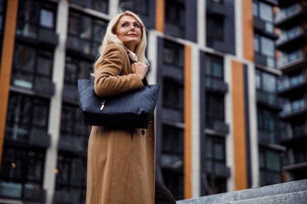Stylish lady standing near an office building Low angle of a calm pleasant female with a handbag on her shoulder looking away older woman purse stock pictures, royalty-free photos & images