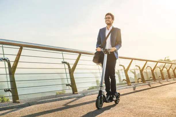 Photo of Ecological transportation concept. Young business man in suit riding electric scooter in office