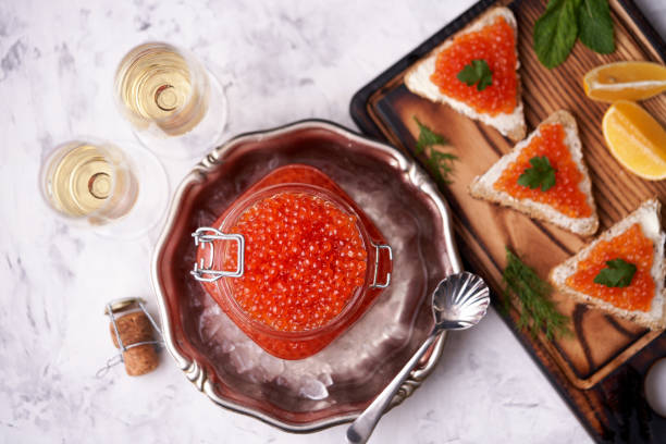 caviar rouge remplissant un bocal en verre au sommet dans une plaque décorative en métal avec de la glace concassée avec une cuillère à côté et deux verres de champagne et sandwichs avec caviar rouge et légumes verts sur un fond en pierre légère. - freshness seafood crushed ice salmon photos et images de collection