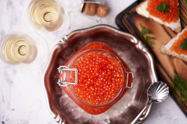 caviar rouge remplissant un bocal en verre au sommet dans une plaque décorative en métal avec de la glace concassée avec une cuillère à côté et deux verres de champagne et sandwichs avec caviar rouge et légumes verts sur un fond en pierre légère. - freshness seafood crushed ice salmon photos et images de collection