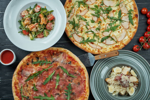 istock Preserved dining table with different dishes: pizza, Caesar salad and seafood salad on a black background. Top view, food flat lay 1216179195