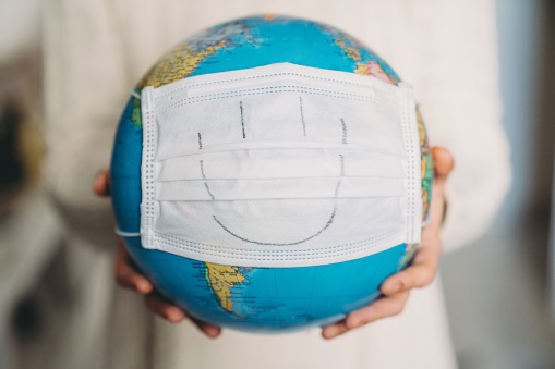 Young woman holding a globe with a face mask on it - Conceptual Coronavirus Covid-19 virus pandemic. A smile is drawn on the face mask.