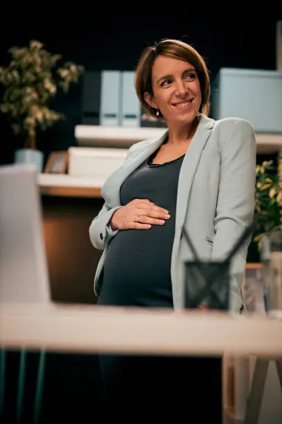 Positive smiling Caucasian pregnant businesswoman standing in home office touching her belly.