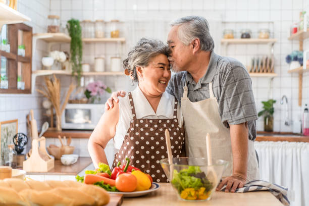 coppia anziana asiatica felice di vivere in cucina. nonno che cucina il piatto di insalata con la nonna con felicità, baciandosi goditi la vita di pensionamento insieme. relazioni e attività degli anziani. - 60s senior adult breakfast cheerful foto e immagini stock