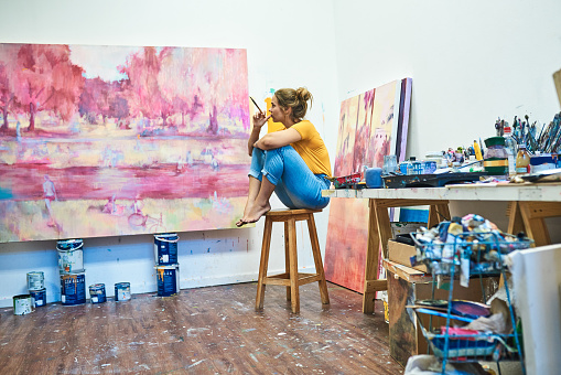 Shot of a young woman sitting on a stool while looking at a painting in an art studio