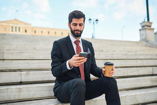 Shot of a young businessman using a cellphone while drinking coffee on a staircase the city