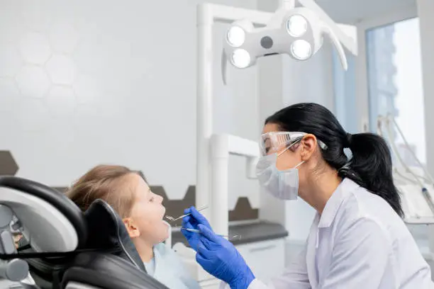 Young female dentist in whitecoat, protective eyeglasses, mask and gloves going to do oral checkup of oral cavity of her little patient