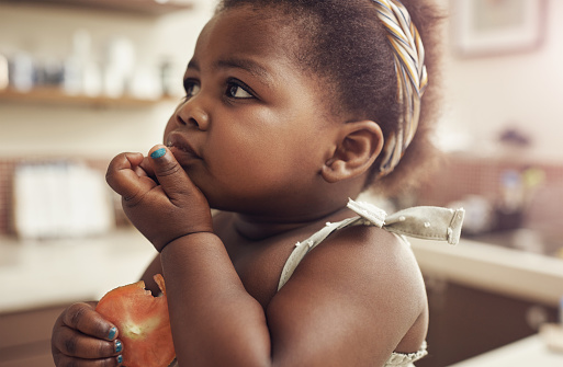 Portrait of an adorable baby girl eating a fresh slice of tomato in the kitchen at home