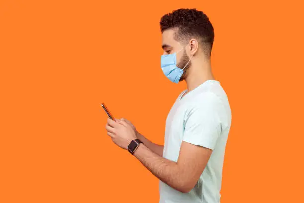 Side view of happy brunette man with surgical medical mask smiling while using cellphone, chatting in social media, enjoying mobile service. empty copy space for advertisement, indoor studio shot