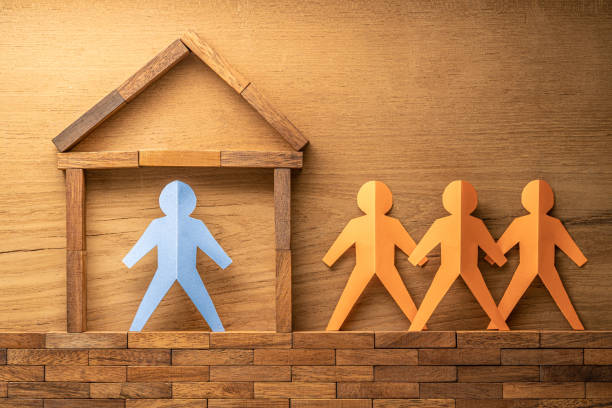 Blue paper cutout human figure inside a wooden block made home and three other orange paper cutout figures outside of the wooden house on wood background, stay at home during COVID-19 virus outbreak concept figura humana recortada de papel azul dentro de un bloque de madera hecho casa y otras tres figuras de recorte de papel naranja fuera de la casa de madera sobre fondo de madera, quédese en casa durante el concepto de brote de virus covid-19