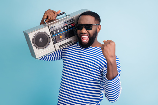 Photo of funny dark skin guy hold retro cassette recorder on shoulder, raise fist amazing students party chill wear sun glasses striped sailor shirt isolated blue color background