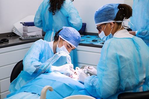 Professional female dentist and assistant in medical masks and caps curing teeth of patient during work in modern dental clinic