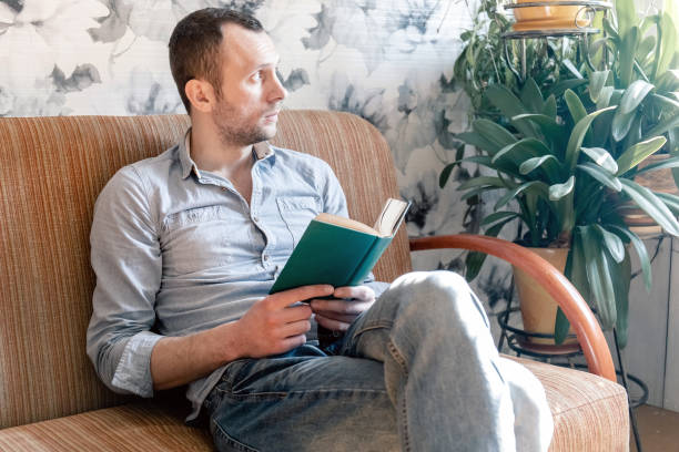 istock young man reading a book while sitting on a sofa and looking sadly to the side, home isolation during quarantine coronovirus covid-19 1216229803
