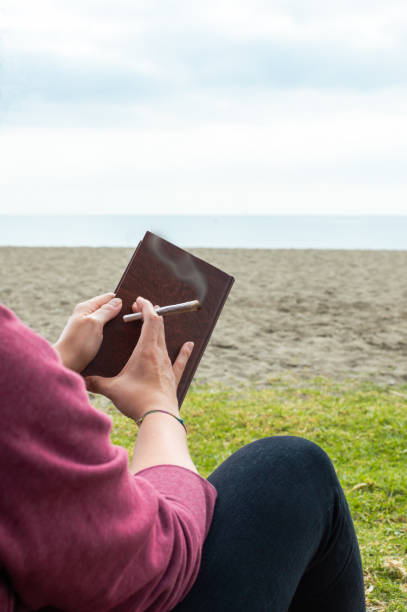 Person reading a book on beach take a break to smoke a marijuana joint or cigarette. Blur background with sea and sky.