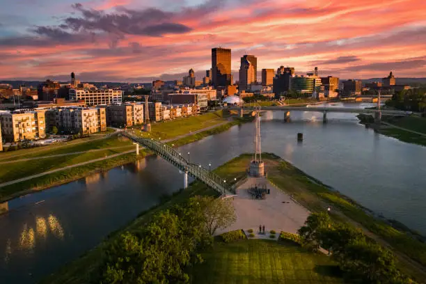 An aerial drone view over a park looking towards downtown Dayton, Ohio at sunset at the confluence of the Great Miami and Mad Rivers with brightly colored clouds in the sky.
