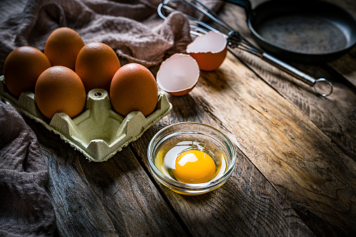 Cooking: eggs in a rustic wooden table. Half dozen eggs are in a cardboard container, one egg is cracked and the egg yolk is in a glass bowl. A wire whisk and a cooking pan are at the top right. The composition is at the left of an horizontal frame leaving useful copy space for text and/or logo at the right. Predominant color is brown. High resolution 42Mp studio digital capture taken with SONY A7rII and Zeiss Batis 40mm F2.0 CF lens