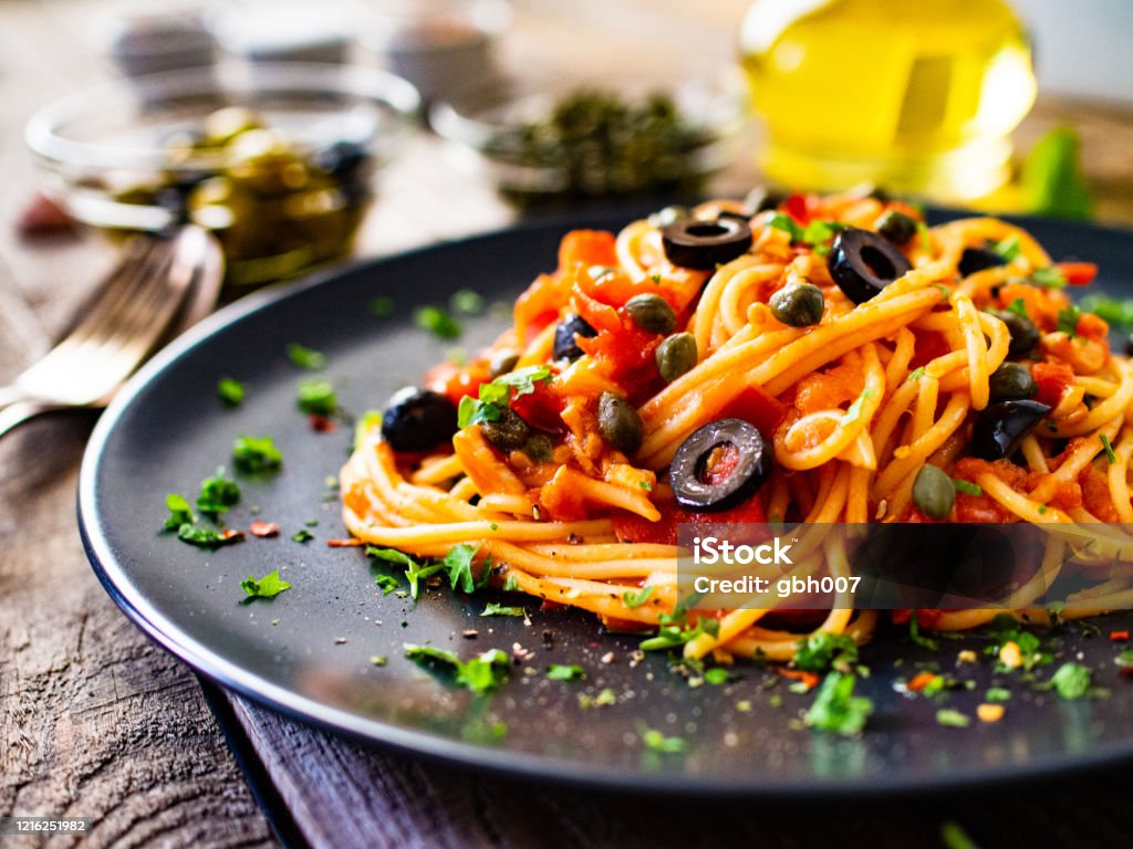 Pasta puttanesca with tomato sauce, anchovies, chilli, capers and olives on wooden table Puttanesca Stock Photo