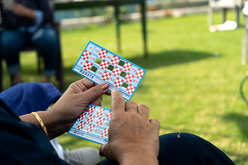 Delhi, India - circa 2020 : Shot with selective focus, showing an old lady's hand holding a tambola bingo card at an outdoor party with others in the background and out of focus. This is a very popular kitty party game in delhi, mumbai india with multiple people playing for cash prizes