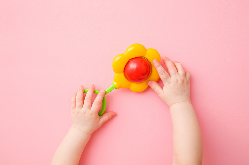 Manos de niña jugando con sonajero de flores de colores en el fondo del suelo de color rosa claro. Color pastel. Closeup. Juguete de desarrollo para lactantes. Vista de arriba hacia abajo. photo