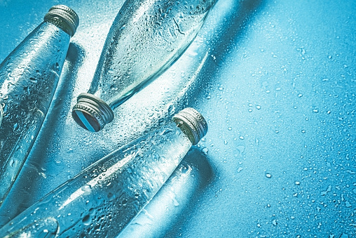 A glass bottle of water on blue background splashes drops of water on top. Quench thirst. Background water drops place and text copy space