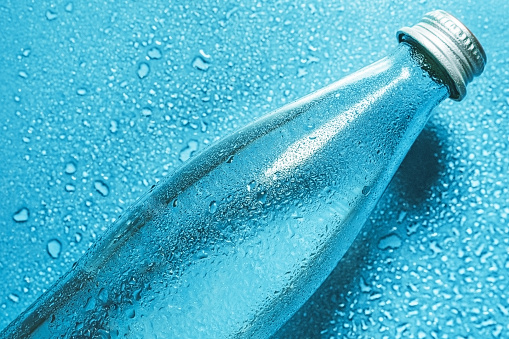 A glass bottle of water on blue background splashes drops of water top. Quench thirst. Background water drops place for text copy space