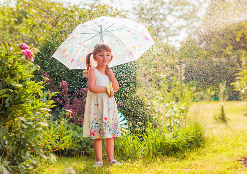 Happy little girl plays in garden under the summer rain with an umbrella.
