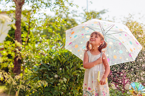 Happy little girl plays in garden under the summer rain with an umbrella. Expressive facial emotion.