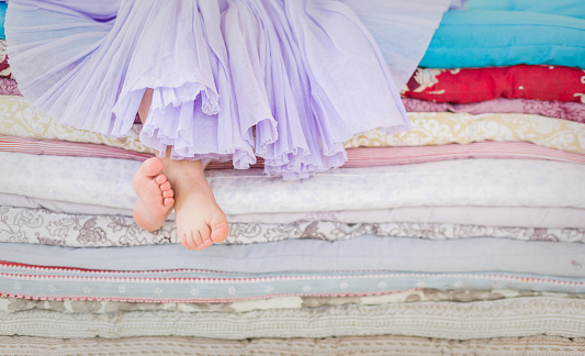 The fairy tale the princess on a pea. Little girl in lilac tutu skirt sitting on the high bed. Bare foot if the girl. Legs of a little girl sitting on a pile of colorful mattresses.