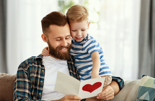 Happy young bearded man in casual wear reading greeting card while sitting with little son on sofa at home in fathers day