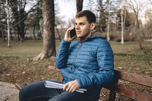 Young blind man having a phone call while sitting on a park bench