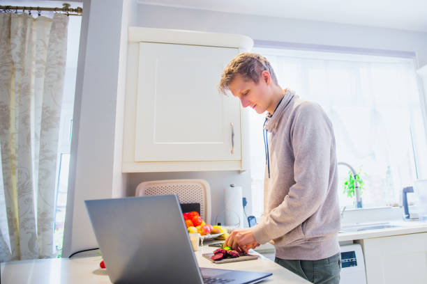 istock Young caucasian man working from home. Freelancer cooking vegetarian lunch on the kitchen table and working on pc laptop. Home office, remote workplace concept. Soft selective focus. Copy space. 1216272254