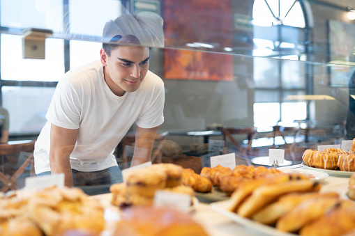 Attractive Hispanic man looking through a glass and choosing some bread and pastries in a bakery shop